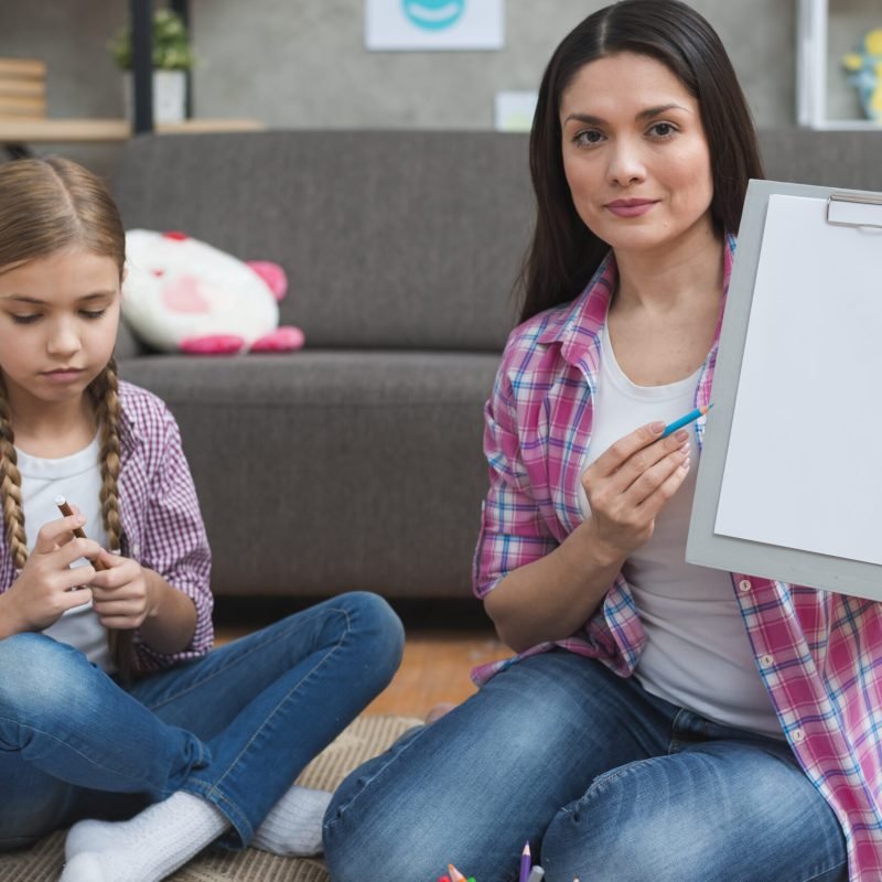 professional-female-psychologist-sitting-with-girl-carpet-showing-white-paper-clipboard